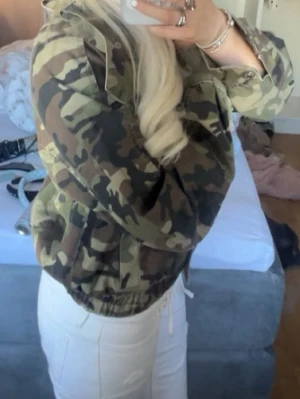 Snygg jacka från Gina - Snygg vindjacka i camouflage-mönster med huva och dragsko i midjan. Jackan har fickor med lock och stängs med dragkedja. Använd 1 gång, helt ny.