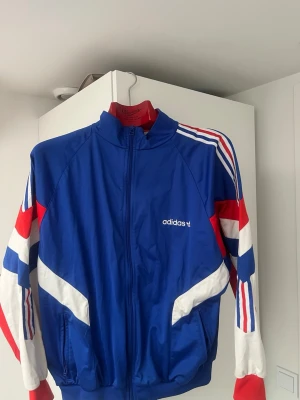 Blå vindjacka från Adidas - Snygg blå vindjacka från Adidas med vita och röda detaljer samt klassiska ränder på ärmarna. Jackan har dragkedja framtill, hög krage och två fickor. Perfekt för en sportig och retro look.