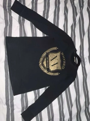 Svart sweatshirt från Whyred - Snygg svart sweatshirt från Whyred med stort guldfärgat tryck framtill. Tröjan har rund halsringning och långa ärmar. Perfekt för dig som gillar stilrena plagg med en twist. Materialet känns mjukt och bekvämt.