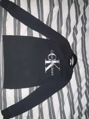 Svart Calvin Klein sweatshirt - Snygg svart sweatshirt från Calvin Klein Jeans med stort silverfärgat CK-logotyptryck på bröstet. Tröjan har rund halsringning och långa ärmar. Perfekt för dig som gillar stilrena och klassiska plagg med en modern touch.