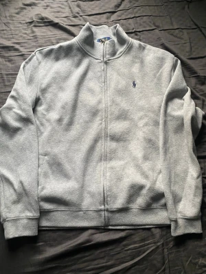 Grå zip hoodie från Polo Ralph Lauren - Snygg grå zip tröja från Polo Ralph Lauren med klassisk broderad logga på bröstet. Tröjan har hög krage, dragkedja hela vägen och två fickor framtill. Perfekt för en chill och stilren look.