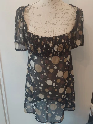 Svart transparent prickig blus - Snygg svart transparent blus med beige och vita prickar i olika storlekar. Blusen har korta puffärmar, rundad urringning och ett band i midjan för knyt. 