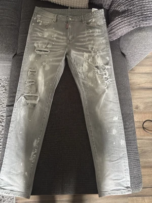 Grå slitna jeans med färgstänk - Grå jeans med raka ben, slitningar och hål på framsidan samt vita färgstänk över hela byxan. Klassisk femficksmodell med knappgylf och coolt, trendigt uttryck. Perfekt för dig som gillar streetstyle och en avslappnad look.