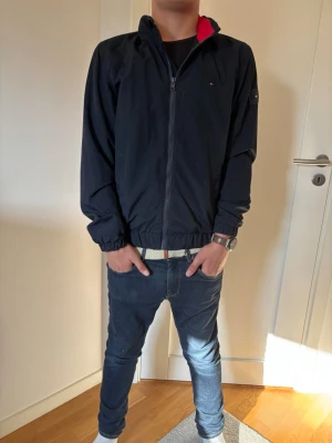 Svart overshirt från Tommy Hilfiger - Snygg svart overshirt från Tommy Hilfiger med dragkedja framtill och diskret logga på bröstet samt ärmen. Jackan har elastiska muddar vid ärmslut och midja, samt röd detalj på insidan av kragen. Perfekt för lager-på-lager-stil.