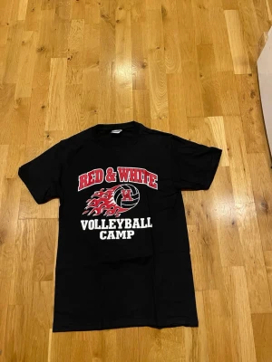 Svart t-shirt Red & White Volleyball stl S  - Cool svart t-shirt med tryck 'Red & White Volleyball Camp' i rött och vitt framtill. Klassisk passform med rund hals och korta ärmar. Perfekt för dig som gillar volleyboll och sportig stil.