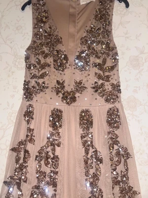 Champagnefärgad långklänning Maya Deluxe - Champagnefärgad långklänning från Maya Deluxe med v-ringning och vackra paljettbroderier i blommigt mönster. Klänningen har ärmlös design och är tillverkad i ett lätt, flowy material som ger en lyxig känsla.