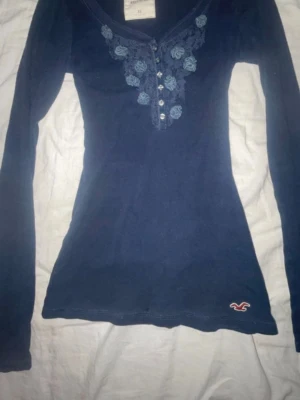 Mörkblå långärmad topp Hollister - Mörkblå långärmad topp från Hollister vintage med broderade blommor och knappar framtill. Slimmad passform och liten logga nertill. Perfekt för dig som gillar detaljer och en klassisk stil.