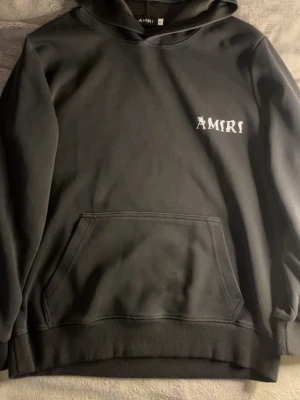 Svart hoodie från Amiri med tryck - Svart hoodie från Amiri med vit logga tryckt på bröstet och stort tryck på ryggen. Klassisk känguruficka framtill, ribbade muddar och huva. Perfekt för en streetwear-look och riktigt skön att bära. Materialet känns mjukt och följsamt.