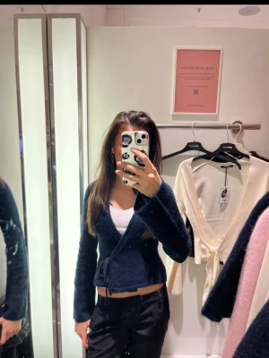 Mörkblå omlottkofta från Gina Tricot - Mysig mörkblå omlottkofta från Gina Tricot med knytband i midjan. Koftan har lång ärm, mjuk och fluffig känsla samt en kortare, figurnära passform. Perfekt att styla över ett linne eller t-shirt för en trendig look.