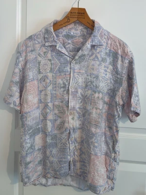 Mönstrad kortärmad skjorta från Zara - Kortärmad skjorta från Zara med pastellfärgat geometriskt mönster i lila, rosa, grön och vit. Skjortan har klassisk krage, knappar framtill och är tillverkad i ett lätt och luftigt material, perfekt för varma dagar.