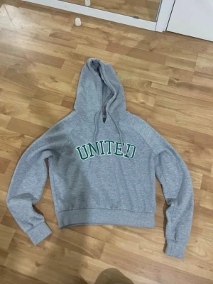 Grå hoodie med UNITED-tryck - Snygg grå hoodie med huva och dragsnören. På bröstet står det UNITED i gröna bokstäver med vit kant. Hoodien har ribbade muddar och är tillverkad i mjukt sweatshirtmaterial. Kan vara lätt nopprig på vissa ställen. Osäker men tror den är från hm way back!