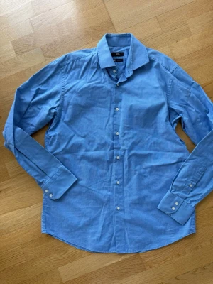 Blå skjorta från BOSS Regular Fit - Snygg blå skjorta från BOSS i regular fit med klassisk krage och vita knappar. 