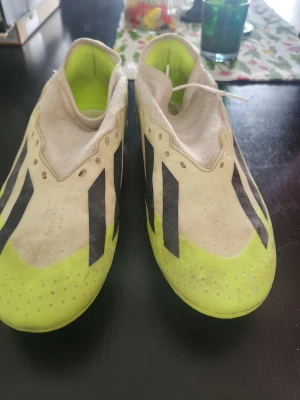 Adidas gula och vita fotbollsskor - Säljer ett par Adidas fotbollsskor i en fräsch gul och vit färg med svarta detaljer. Skorna har låg profil, klassiska tre svarta ränder och är tillverkade i syntetmaterial med gummidobbar för bra grepp på planen. Perfekta för dig som vill sticka ut på matchen!