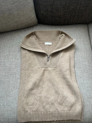 Beige stickad väst med halvzip H&M - Beige stickad väst från H&M med halvzip och hög krage. Ribbstickade detaljer vid krage, ärmhål och nederkant. Perfekt att slänga över en skjorta eller t-shirt för en snygg lager-på-lager-look.