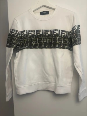 Vit Fendi sweatshirt - Snygg vit sweatshirt från Fendi med ett brett band över bröstet och armarna med klassiskt Fendi-logomönster i svart och grått.