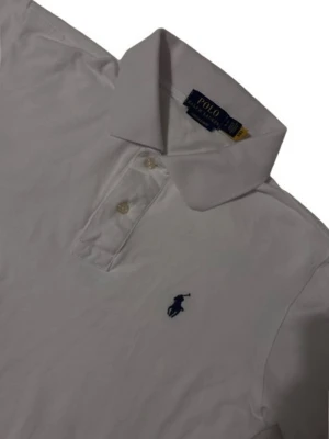 Vit pikétröja från Polo Ralph Lauren - Klassisk vit pikétröja från Polo Ralph Lauren med broderad marinblå logga på bröstet. Tröjan har krage, korta ärmar och knäppning med två knappar framtill. Tillverkad i mjuk bomull som känns skön mot huden. Vit men ses lite grå ut i bilden