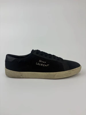 Svarta Saint Laurent sneakers - Svarta sneakers från Saint Laurent med handskriven logga på sidan. Skorna har en klassisk låg profil, svarta skosnören och vit sula med vintagekänsla. Tillverkade i läder och textil för en stilren och tidlös look.