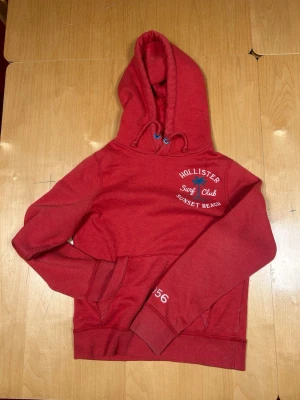 Röd hoodie från Hollister - Röd hoodie från Hollister med tryck 'Surf Club Sunset Beach' och palm på bröstet. Klassisk känguruficka framtill, ribbade muddar och dragsko i huvan. Siffrorna 1956 och 19 tryckta på ärm och bak. Perfekt för en avslappnad stil.