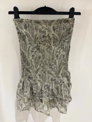 Grön kort klänning med volang - Säljer en beige kort klänning från H&M med smockad överdel och volang nedtill. Klänningen är axelbandslös och har ett diskret paisleymönster i grå toner. Perfekt för sommarens alla tillfällen!