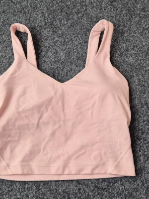 Ljusrosa linnetopp från Lululemon - Superfin ljusrosa linnetopp från Lululemon med v-ringning och breda axelband. Toppen är croppad och tillverkad i ett mjukt, stretchigt material som passar perfekt till träning eller chill. Enkel och stilren design med liten logga på ryggen.
