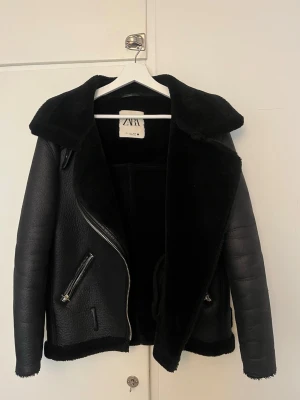 Svart pälsjacka från Zara - Snygg svart pälsjacka från Zara med bred krage och dragkedjor i silver. Jackan har en klassisk bikerstil med fuskpäls på insidan och detaljerade sömmar. Perfekt för att hålla värmen under kalla dagar och ger en cool look.
