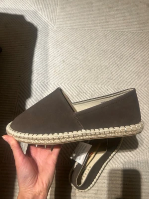 Bruna loafers i mocka med flätad sula - Snygga bruna loafers i mocka med klassisk espadrille-inspirerad flätad sula. Skorna har en stilren och enkel design utan snörning, perfekt för en avslappnad look. Materialet är mjukt och ger en skön känsla på foten.