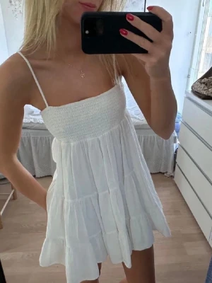 Vit sommarklänning  - Supersöt vit kort klänning med tunna axelband och smockad överdel. Klänningen har volanger och är tillverkad i ett lätt bomullstyg, perfekt för varma dagar. Den har en luftig passform och är ärmlös för en avslappnad och fräsch look. Slutsåld!! 💗💗