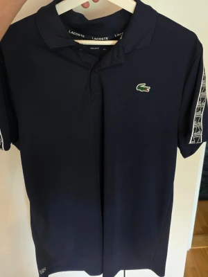 Mörkblå pikétröja från Lacoste - Mörkblå pikétröja från Lacoste med klassisk krage och knäppning framtill. Tröjan har vita mönstrade detaljer på ärmarna och den ikoniska gröna krokodilloggan på bröstet. Perfekt för en sportig och stilren look.