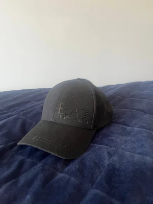 Svart keps från EA7 Emporio Armani - Snygg svart keps från EA7 Emporio Armani. Klassisk böjd skärm och justerbar rem bak. Skick 10/10 endast testad. Storlek M. Nypris 600kr+.