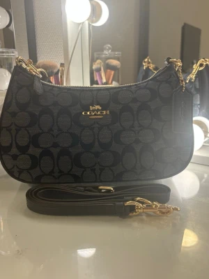 Coach väska - Snygg svart handväska från Coach med klassiskt monogrammönster och guldfärgade detaljer. Väskan har en avtagbar axelrem i läder och dragkedja upptill. Perfekt för dig som gillar stilrena accessoarer med lyxig känsla.