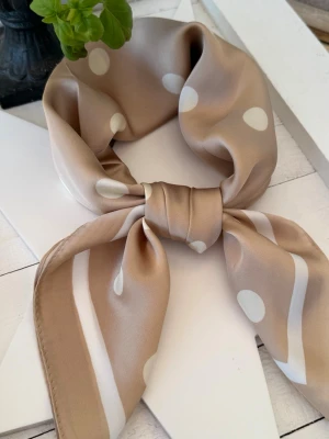 Latte Beige Polka Dot Scarf – silkeslook & old money chic  - Elegant scarf i mjuk latte beige ton med klassiska polkaprickar och exklusiv satinfinish. Den lena silkeslooken ger ett sofistikerat fall och en tidlös “old money”-känsla med modern couture-touch.  Vackert neutral färgpalett i creamy beige och soft ivory som passar året runt – lika chic runt halsen som knuten på väskan eller i håret.  Lätt, feminin och lyxig accessoar med exklusiv vintageinspirerad estetik.
