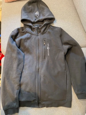 Svart Sail Racing hoodie med dragkedja - Svart hoodie från Sail Racing med hel dragkedja framtill och bröstficka med dragkedja. Märkets logga syns på bröstet och på huvan. Hoodien har ribbade muddar och är tillverkad i mjukt material, perfekt för en avslappnad stil.