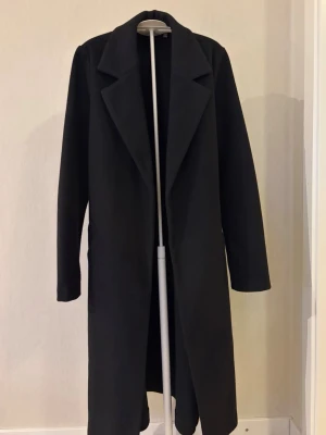 Svart lång kappa med skärp - Jag säljer en svart lång kappa med knytning i midjan. Kappan är från Vero Moda i storlek XS. Är använd ett fåtal gånger och har inga defekter.