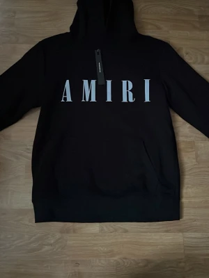 Amiri hoddie  - Svart hoodie från Amiri med stor vit logga framtill. Klassisk modell med huva och känguruficka. Mjuk bomullskänsla och ribbade muddar vid ärmslut och nederkant. Perfekt för en streetwear-look.