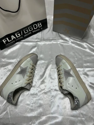 Golden Goose Superstar sneakers glitter - Snygga vita Golden Goose Superstar sneakers med grå stjärna på sidan och silverglittrig häl. Skorna har beige mockadetaljer vid tån och klassiska vita skosnören. Ovandelen är i läder och sulan är beige. Perfekta för dig som vill sticka ut med stil.