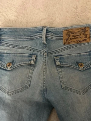 Ljusblå jeans från Deisel - Ljusblå jeans från deisel med bootcut/rak-passform och klassiska fem fickor. Jeansen har slitningar och en snygg tvättad look, samt detaljerade bakfickor med knapp. Perfekta för en avslappnad och trendig stil.