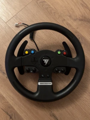 Thrustmaster ratt till spelkonsol - Säljer en svart Thrustmaster ratt med klassisk rund form och flera knappar i olika färger. Ratten har paddlar för växling och är tillverkad i plast och konstläder. Perfekt för racing- och bilspel på konsol.