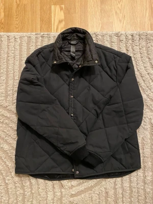 Svart Chrome Hearts quiltad jacka med tryckknappar - Snygg svart Chrome Hearts quiltad jacka med hög krage och tryckknappar framtill. Jackan har ett diskret rutmönster, ribbade muddar vid ärmsluten och en stilren look. Perfekt för kyliga dagar och enkel att matcha med olika outfits.