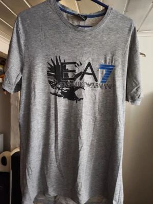 Grå EA7 Emporio Armani t-shirt Helg REA NU! - Snygg grå t-shirt från EA7 Emporio Armani med stort tryck framtill i svart och blått. T-shirten har rund hals, korta ärmar och är gjord i mjukt bomullsmaterial. Perfekt för en clean och stilren look.