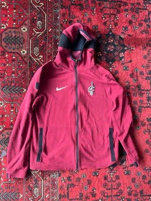 Vinröd Nike Cavaliers hoodie - Snygg vinröd hoodie från Nike med Cleveland Cavaliers-logga på bröstet och dragkedja framtill. Huva med svart innerfoder och två svarta fickor på sidorna. Perfekt för dig som gillar basket och vill ha en sportig stil.