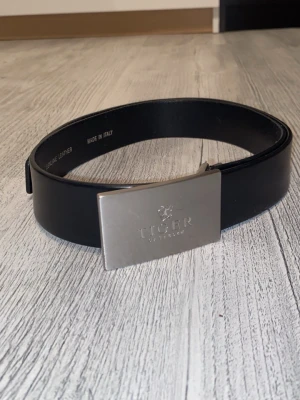 Svart skärp från Tiger of Sweden (85 cm) - Snyggt svart bälte från Tiger of Sweden med stilren, rektangulär silverspänne. Tillverkat i äkta skinn och har en clean look som passar till många outfits. Bältet är tillverkat i Italien och har diskret logga på spännet.