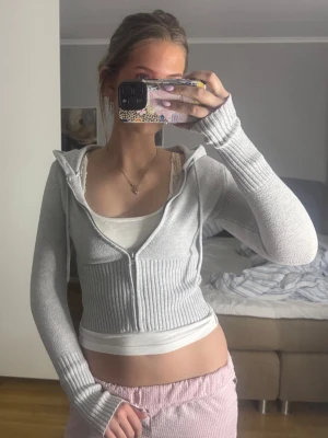 Grå ribbad hoodie med dragkedja - Säljer en grå, ribbad hoodie med dragkedja och huva. Modellen är croppad med långa ärmar och tumhål. Stickad känsla och snören i huvan. Perfekt för dig som gillar en chill och trendig look.