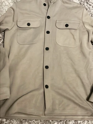 Beige overshirt från Only & Sons - Stilren beige overshirt från Only & Sons med svarta knappar och två stora bröstfickor med lock. Skjortjackan har rak passform och är tillverkad i ett mjukt tyg som känns bekvämt. Perfekt att bära öppen eller stängd över en t-shirt.