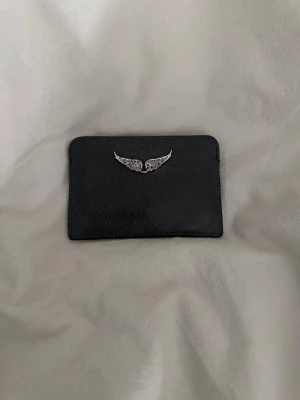 Zadig korthållare med silvervingar - Snygg svart korthållare från Zadig&Voltaire. Perfekt för dig som vill ha ett stilrent och unikt accessoar till dina kort. Enkel och smidig att ha i fickan eller väskan.🌟