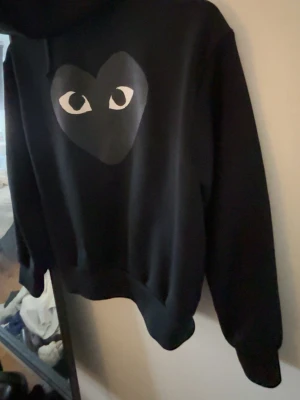 Svart Comme des Garçons Play hoodie - Säljer en svart hoodie från Comme des Garçons Play med det ikoniska hjärtat med ögon tryckt på bröstet. Hoodien har dragkedja, fickor framtill och en klassisk huva. Perfekt för dig som gillar streetwear och vill ha något stilrent men ändå unikt.