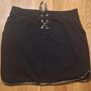 Svart kortkjol med snörning - Svart kortkjol med snörning framtill och elastisk midja. Kjolen har fickor på sidorna och en kontrasterande kant längst ner. Perfekt för en avslappnad och sportig stil.