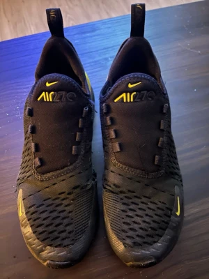 Nike Air 270 svarta/gula sneakers - Säljer ett par Nike Air 270 sneakers i svart med gula detaljer. Skorna har meshmaterial på ovandelen för bra ventilation och en tydlig Air-sula bak för extra komfort. Swoosh-logga och AIR270-tryck i gult ger en sportig vibe. Perfekta för dig som gillar streetwear och snygg design.