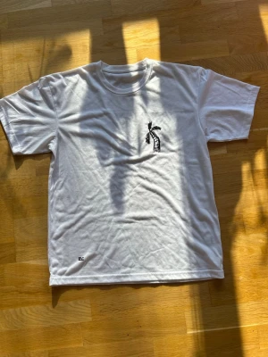 Vit t-shirt med palmtryck  - Vit t-shirt med ett litet svart palmtryck på bröstet. Klassisk rund halsringning och korta ärmar. T-shirten har en clean look, perfekt för en avslappnad stil.
