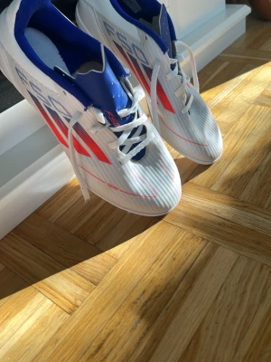 Adidas vita fotbollsskor med röd detalj - Snygga fotbollsskor från Adidas i vitt med röda och blå detaljer. Skorna har en strömlinjeformad design, vita snören och ovandel i syntetmaterial för bra bollkontroll. Perfekta för dig som vill sticka ut på planen.