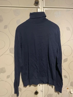 Mörkblå polotröja i stickat - Mörkblå stickad polotröja med lång ärm och ribbade muddar vid ärmslut och nederkant. Klassisk och enkel design, perfekt för lager-på-lager. Hög polokrage som ger extra värme. Materialet känns mjukt och följsamt.